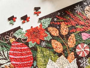 A close-up of a completed jigsaw puzzle showing a colorful holiday design with red ornaments, green leaves, orange flowers, and white snowflakes, with a few loose puzzle pieces on a light surface.