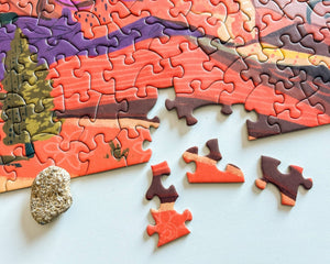 A partially completed landscape jigsaw puzzle with loose pieces scattered on a light surface, including a small stone prop.