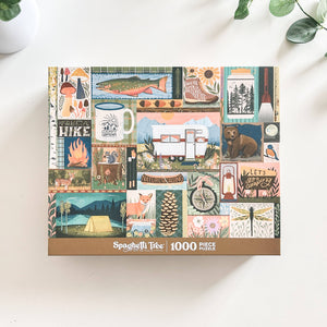 A 1000-piece jigsaw puzzle box by Spaghetti Tree, featuring a colorful collage of outdoor camping and nature illustrations, photographed on a white surface with a plant in the background.