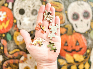 A hand holds several small, random cut puzzle pieces with an autumn and Halloween-theme, set against a festive, out-of-focus shot of a partial view of the completed jigsaw puzzle.