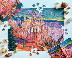 A colorful jigsaw puzzle showing a canyon landscape, arranged on a white surface with loose puzzle pieces, stones and the puzzle box around it. A poster of puzzle design appears in the lower right corner.
