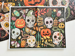 An art print featuring a colorful Halloween design with sugar skulls, jack-o'-lanterns, candelabras, and butterflies on a black background, displayed on a white surface with a partial view of a completed puzzle featuring the same art.