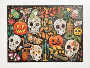 A completed jigsaw puzzle featuring a colorful Halloween collage with sugar skulls, jack-o'-lanterns, butterflies, leaves, candles, and spiders on a dark background, photographed against a plain white backdrop.