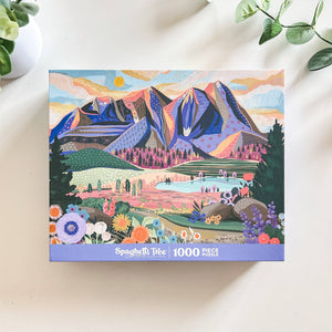 Top view of puzzle box showing illustration of beautiful mountain and valley scene. Box is on white background surrounded by small foliage.