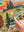 A close-up of a completed landscape jigsaw puzzle showing a colorful mountain valley scene with trees, a river, bison, and a corner of the rainbow layers of a hot spring. A person's finger pushes a piece towards a empty section of the puzzle. 
