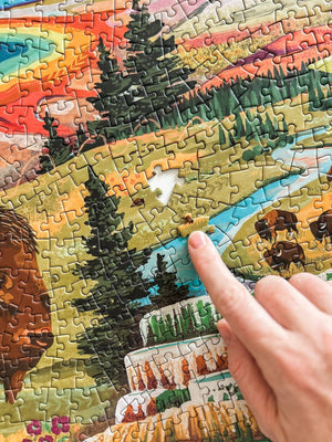 A close-up of a completed landscape jigsaw puzzle showing a colorful mountain valley scene with trees, a river, bison, and a corner of the rainbow layers of a hot spring. A person's finger pushes a piece towards a empty section of the puzzle. 