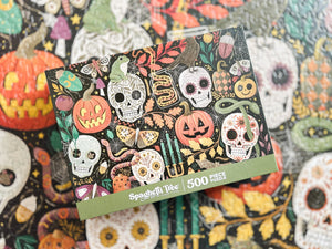 A 500-piece jigsaw puzzle box by Spaghetti Tree, featuring a colorful Day of the Dead/Halloween illustration with sugar skulls, jack-o'-lanterns, butterflies, and candles, shown resting on a matching patterned puzzle surface.