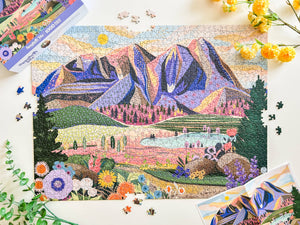 Almost complete puzzle showing lovely valley and mountain scene on white table surrounded by flowers, puzzle box and puzzle poster. 