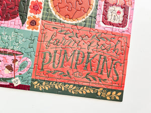 A close-up of a completed jigsaw puzzle featuring a fall harvest design with the words 'farm fresh PUMPKINS' in dark green script on a coral background, surrounded by floral and pumpkin illustrations in red, pink, teal, and gold.