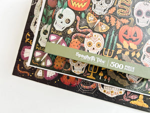 A close-up of a 500-piece jigsaw puzzle box by Spaghetti Tree with a Halloween-themed design featuring sugar skulls, pumpkins, spiders, and candles on a dark background.