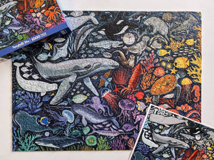 Completed jigsaw puzzle with ocean-themed design featuring whales and sea creatures on white surface with box lid and poster of puzzle.