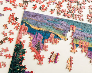 A partially completed jigsaw puzzle showing a colorful mountain and canyon landscape with pink, purple, blue, green, and yellow tones, surrounded by scattered puzzle pieces on a white surface.