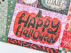 A close-up of a completed jigsaw puzzle featuring a large orange pumpkin with the words “Happy Halloween” in black lettering, surrounded by layered autumn and Halloween-themed puzzle pieces.