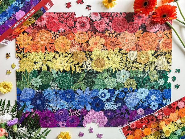 Colorful floral rainbow puzzle with a partially completed design on a white surface surrounded by flowers, puzzle box lid and poster.