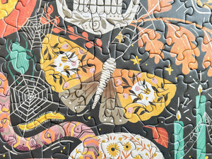 A close-up of a completed jigsaw puzzle featuring a colorful Halloween design with a large butterfly, spiderwebs, candles, and floral motifs on a dark background.
