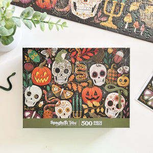 A 500-piece jigsaw puzzle box by Spaghetti Tree, featuring a colorful Day of the Dead/Halloween design with skulls, pumpkins, snakes, and candles on a black background. The box is displayed on a white desk with a plant and other puzzle pieces nearby.