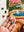 A hand holds a random-cut jigsaw puzzle piece with a rainbow pattern in front of a partially completed colorful puzzle.