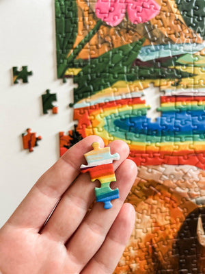 A hand holds a random-cut jigsaw puzzle piece with a rainbow pattern in front of a partially completed colorful puzzle.