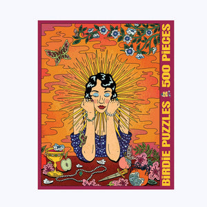 Photo of a jigsaw puzzle box against a white surface/wall. Box shows an illustration of a beautiful woman with short black hair and an array of spikes radiating from her head, surrounded by flowers, a knife, apple and a few other small items. 