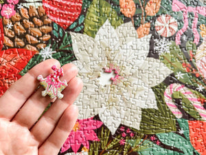 A hand holds a small, colorful jigsaw puzzle piece featuring a pink and white floral design, set against a background of a jigsaw puzzle with a winter holiday theme.