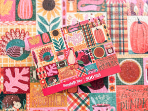 A 500-piece jigsaw puzzle box by Spaghetti Trees featuring a colorful autumn patchwork design with pumpkins, corn, leaves, and plaid patterns, displayed on a out of focus shot of a completed version of that puzzle in the background.