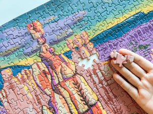 A close-up of a colorful jigsaw puzzle showing an abstract landscape or canyon scene, with a hand holding a small pink puzzle piece over the blank white gap in the puzzle.